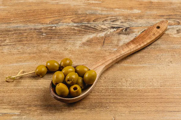 Ahşap bir kaşık, zengin renklerini ve dokusunu kırsal bir ahşap arka planda sergileyen birkaç yeşil zeytin barındırır. Yemek sunumlarını ve yemekleri geliştirmek için mükemmel..