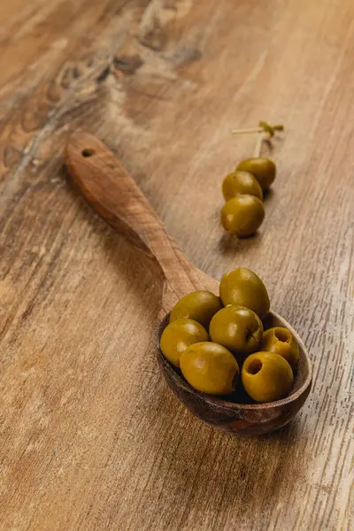 Kırsal bir ahşap kaşık, doğal ve rahat bir atmosferi çağrıştıran ahşap bir yüzeye yerleştirilmiş birkaç yeşil zeytin tutar. Yakınlarda, daha fazla zeytin serpilmiş..