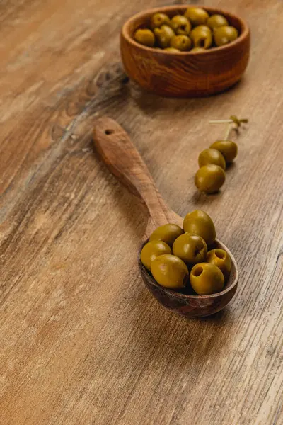 Yeşil zeytinler ahşap bir kaşığa ve kırsal bir ahşap masada bir kaseye konur. Doğal ışık sıcak bir atmosfer yaratıyor, odunların ve zeytinlerin dokusunu vurguluyor..