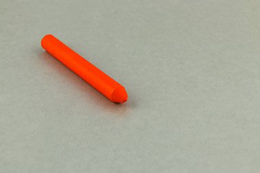 A bright orange crayon lies on a smooth gray surface. Natural light highlights its vibrant color, offering a pop against the neutral background.