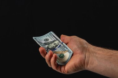 A hand presents a one hundred dollar bill, showcasing the detailed design. The background is dark, emphasizing the money's vibrant colors.