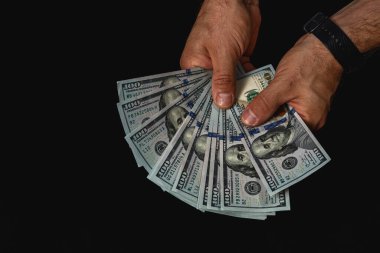 Two hands hold a fan of hundred dollar bills, showcasing the currency in dim lighting. The focus highlights the details of the bills and the texture of the hands.