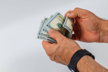 A person is counting several hundred dollar bills with both hands while wearing a black wristwatch. The setting has a simple, neutral background.