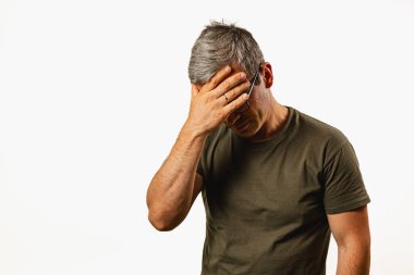 A middle-aged man stands in an indoor setting, showing visible signs of frustration with his hand covering his forehead. His expression reflects deep thought or distress.