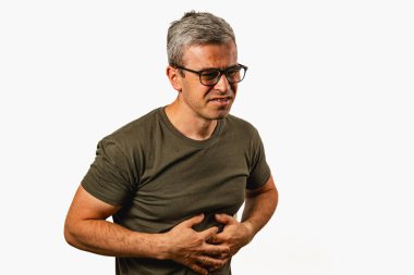 A middle-aged man stands at home, holding his stomach with a pained expression. He wears glasses and a green shirt, indicating discomfort or illness.