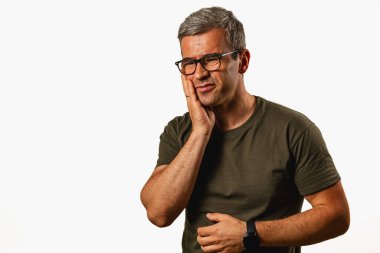 A middle-aged man stands indoors, showing a thoughtful expression. He gently touches his cheek, suggesting contemplation or concern, dressed casually in a t-shirt.