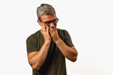 A man stands with a thoughtful expression, gently touching his face. He appears contemplative or distressed, dressed casually in a simple t-shirt, against a neutral background.