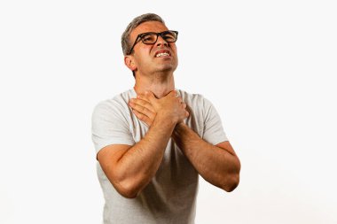 A man in a grey shirt clutches his throat with both hands, expressing discomfort while standing against a plain background. His facial expression shows concern and unease.