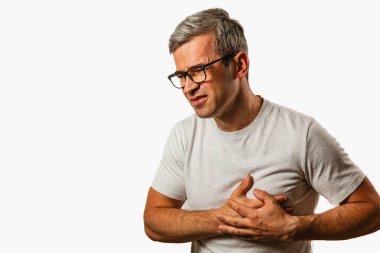 A middle-aged man stands in a plain environment, clutching his chest in visible discomfort, indicating he may be experiencing health issues.