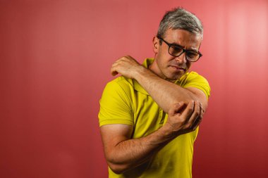A man in a bright yellow shirt is standing against a pink background, rubbing his elbow with a concerned expression as he checks for discomfort in his arm.
