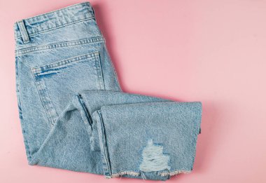 Açık mavi yırtık kot pantolon, düzgün bir şekilde katlanmış pembe bir yüzey üzerinde, gündelik moda ve modaya uygun görünümlü modern kıyafetler sergiliyor..