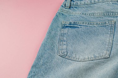 Açık mavi kot pantolonun arka cebinin yakın plan görüntüsü yumuşak pembe bir arka fona yerleştirilmiş, dokuyu renklendiriyor ve kumaşın detaylarını dikiyor..