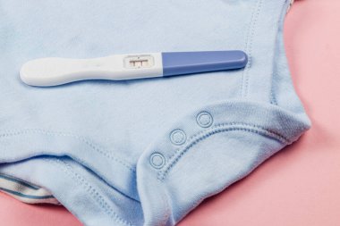 A positive pregnancy test lies next to a light blue baby outfit on a soft pink surface, suggesting joyful anticipation of new beginnings in family life.