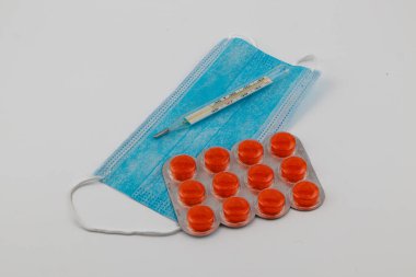 A collection of essential medical supplies is arranged on a white surface. The items include orange tablets, a thermometer, and a light blue face mask.