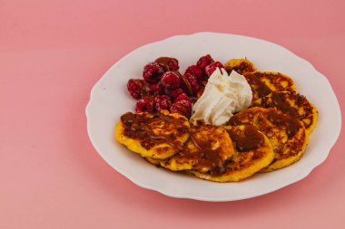 Yumuşak krepler karamel yağmuru, taze ahududu ve kremayla servis edilir. Pembe arka planda nefis bir kahvaltı sahnesi yaratır..