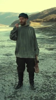 Retro tarzı sakallı balıkçı bir dağ nehrinin kıyısında pipo içiyor.
