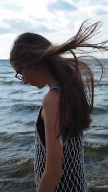 Uzun kahverengi saçlı, gözlüklü ve net elbiseli genç kız kıyı dalgalarında geziniyor.