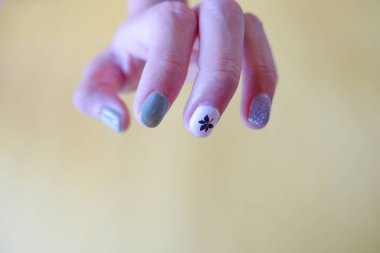 Sarı arka planda güzel manikürlü eller. Jel cilasıyla kaplı genç bir kadının tırnakları. Adaçayı yeşili, çiçek deseni ve parıltılı tırnak cilası..