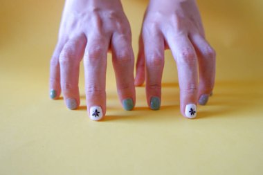 Sarı arka planda güzel manikürlü eller. Jel cilasıyla kaplı genç bir kadının tırnakları. Adaçayı yeşili, çiçek deseni ve parıltılı tırnak cilası..