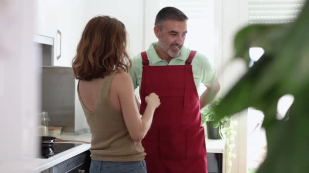キッチンで大人の夫婦と結婚しました 妻は夫が話して笑顔でエプロンを着るのを助けます — ストック動画