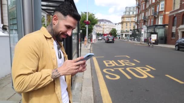 Привлекательный Молодой Мужчина Ждет Автобусной Остановке Оглядываясь Своим Мобильным Телефоном — стоковое видео