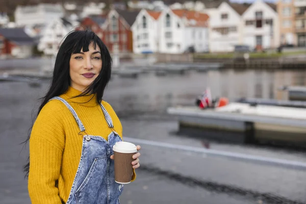 young and beautiful woman with her coffee in her hand looking at the camera with the port in the background and colorful houses out of focus in a village in Norway