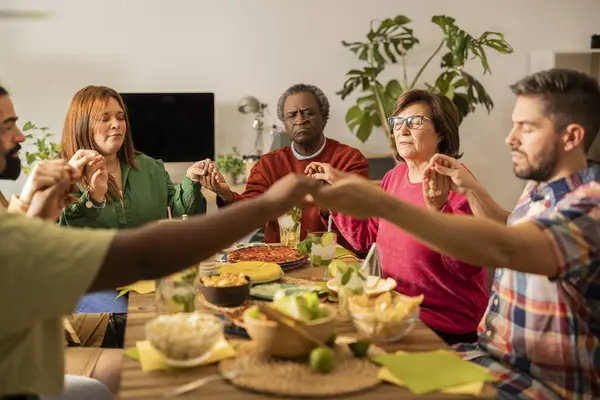 group of multiracial people gathered at home praying before dinner
