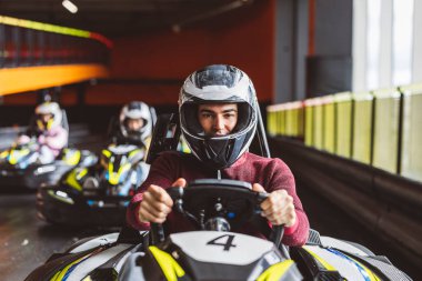 Genç adam kart sürücüsü direksiyonu elinde tutarak yarış arabasındaki kameraya bakıyor.