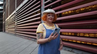 Gülümseyen olgun bir kadın cep telefonuna bakarak sokakta yürüyor.