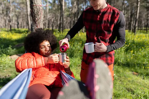 couple in the forest man takes care of his girlfriend by serving her hot tea while she is lying in the hammock