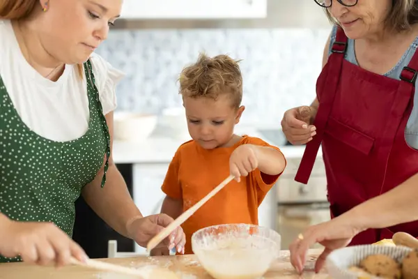 boy cooking with his mother and grandmother learning to help at home holding a wooden spoon