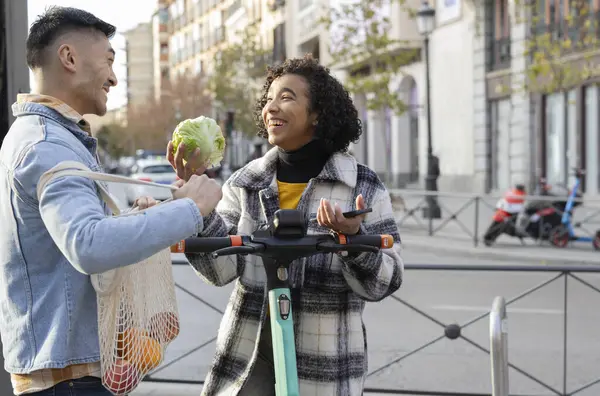 Çevre dostu bir çift sokakta gülüp şakalaşıyor. Kadın elektrikli scooter kullanıyor ve adam çantaları geri dönüştürüyor..