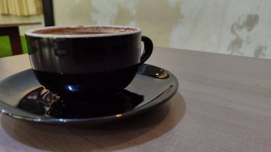 Tahta bir masanın üzerinde kalp şeklinde latte sanat eserleri olan bir fincan sıcak çikolatanın üst görüntüsü. Siyah seramik bir fincan ve çay tabağında sıcak bir içecek..