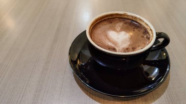 Tahta bir masanın üzerinde kalp şeklinde latte sanat eserleri olan bir fincan sıcak çikolatanın üst görüntüsü. Siyah seramik bir fincan ve çay tabağında sıcak bir içecek..