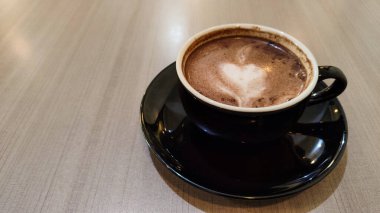 Tahta bir masanın üzerinde kalp şeklinde latte sanat eserleri olan bir fincan sıcak çikolatanın üst görüntüsü. Siyah seramik bir fincan ve çay tabağında sıcak bir içecek..