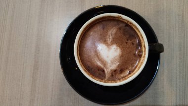 Tahta bir masanın üzerinde kalp şeklinde latte sanat eserleri olan bir fincan sıcak çikolatanın üst görüntüsü. Siyah seramik bir fincan ve çay tabağında sıcak bir içecek..