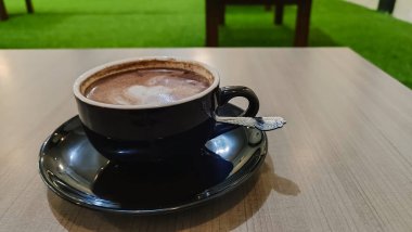 Tahta bir masanın üzerinde kalp şeklinde latte sanat eserleri olan bir fincan sıcak çikolatanın üst görüntüsü. Siyah seramik bir fincan ve çay tabağında sıcak bir içecek..