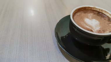 Tahta bir masanın üzerinde kalp şeklinde latte sanat eserleri olan bir fincan sıcak çikolatanın üst görüntüsü. Siyah seramik bir fincan ve çay tabağında sıcak bir içecek..