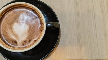 Tahta bir masanın üzerinde kalp şeklinde latte sanat eserleri olan bir fincan sıcak çikolatanın üst görüntüsü. Siyah seramik bir fincan ve çay tabağında sıcak bir içecek..
