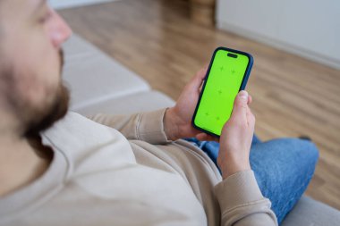  Evdeki adam kanepede uzanmış yeşil model ekranlı akıllı telefon kullanıyor.