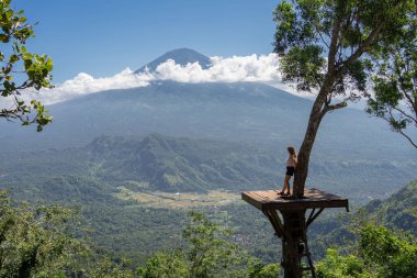 Doğanın tadını çıkaran kadın turist, ağaçtaki fotoğraf noktasında dururken dağlara ve Agung volkanına bakıyor. Bali, Endonezya. Seyahat kavramı