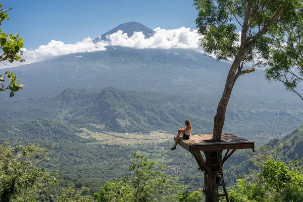 Doğanın tadını çıkaran kadın turist, ağaçtaki fotoğraf noktasında dururken dağlara ve Agung volkanına bakıyor. Bali, Endonezya. Seyahat kavramı