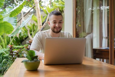 Serbest meslek kavramı. Genç sakallı adam yaz terasında otururken dizüstü bilgisayar kullanıyor. Genç erkek serbest çalışan dışarıda oturuyor ve dizüstü bilgisayarda çalışıyor.