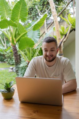 Serbest meslek kavramı. Genç sakallı adam yaz terasında otururken dizüstü bilgisayar kullanıyor. Genç erkek serbest çalışan dışarıda oturuyor ve dizüstü bilgisayarda çalışıyor.