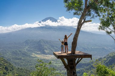 Anne ve kız turistler Endonezya 'daki Bali adasını geziyorlar. Agung Dağı manzaralı ağacın üzerinde dururlar.