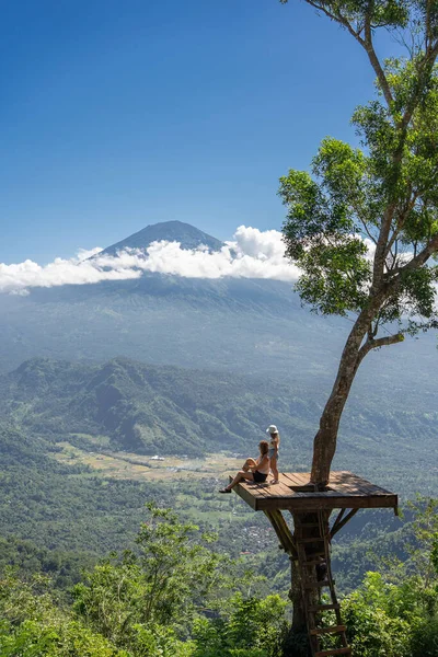 インドネシアのバリ島で旅行を楽しむ母と娘の観光客 彼らはアグン山を眺めながら木の上の写真の場所に立っています — ストック写真