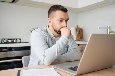 Düşünceli genç beyaz adam mutfak masasında oturmuş dizüstü bilgisayarla düşünüyor. Düşünceli milenyum erkeği, modern aletteki bilgisayar ekranına kablosuz internete bakıyor.