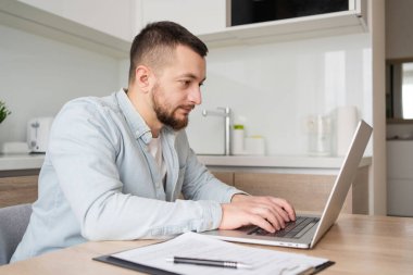 Düşünceli genç beyaz adam mutfak masasında oturmuş dizüstü bilgisayarla düşünüyor. Düşünceli milenyum erkeği, modern aletteki bilgisayar ekranına kablosuz internete bakıyor.