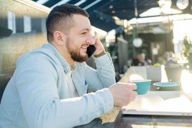 Genç adam bir kafede çalışırken bilgisayar ve telefon kullanıyor.