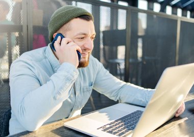 Hipster adam kafede oturmuş bilgisayarla uğraşırken cep telefonuyla konuşuyor. Bir kafede oturmuş dizüstü bilgisayarla çalışan gülümseyen bir iş adamı.